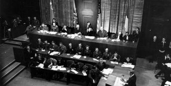 In the first session of the International Military Tribunal in the Palace of Justice in Nuremberg, Germany, Sydney S. Alderman (standing) of the U.S. prosecution panel reads the indictment which charges Hitler's former henchmen with war crimes. The photo shows the presiding judges in the background, sitting before the flags of Russia, Great Britain, the United States, and France