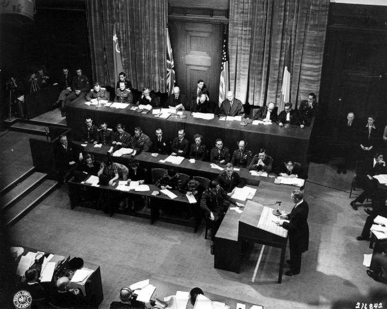 In the first session of the International Military Tribunal in the Palace of Justice in Nuremberg, Germany, Sydney S. Alderman (standing) of the U.S. prosecution panel reads the indictment which charges Hitler's former henchmen with war crimes. The photo shows the presiding judges in the background, sitting before the flags of Russia, Great Britain, the United States, and France