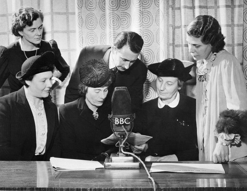 Alors que les employés hommes de la British Broadcasting Company entrent dans les forces armées, les femmes prennent à la BBC les postes de producteur, rédacteur en chef, présentateur, annonceur, créateur d’effets sonores et assurent une part importante et croissante de l’ensemble des émissions de divertissement. Personnes participant à la diffusion de la BBC à Londres, le 15 janvier 1943, pendant Answering you, émission hebdomadaire diffusée sur les ondes courtes aux États-Unis et au Canada: (de gauche à droite) Mme Mary Adams, productrice; Colin Wills, présentateur; et Mme Archer, femme au foyer et travailleuse de guerre; (assis de gauche à droite) Mme Gardner, intendante de la garderie Brook Green; Mme Margaret Biddle, épouse de l’ambassadeur américain auprès des gouvernements alliés à Londres, et Mme Florence Horsbrugh, membre du Parlement, secrétaire parlementaire du ministre de la Santé.